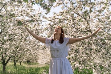 Fantasy woman in long white elegant fashion long dress walks in green spring blossom cherry garden. Happy cheerful girl princess bride. Skirt fabric flies flowing waving in wind motion.