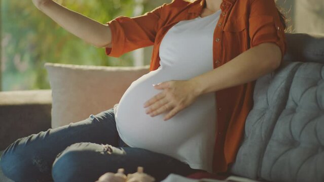 Happy pregnant woman sitting on sofa in home using mobile phone for video call, showing her tummy to relatives