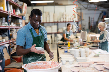 African american worker in pottery factory dips clay plates and cups into a container with glaze