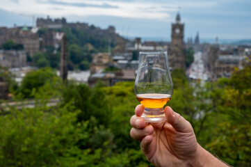 Hand holding glass of single malt scotch whisky and view from Calton hill to park and old parts of Edinburgh city in rainy day, Scotland, UK