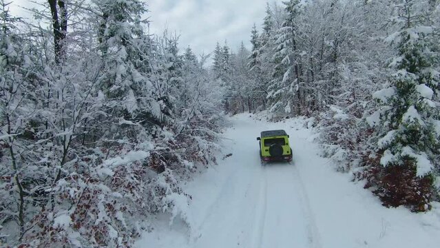 MARCH 2022, LJUBLJANA, SLOVENIA: AERIAL: Amazing performance on Jimny Suzuki 4x4 car driving through snowy forest. Mini 4-wheel drive SUV moving through woodland along snowy winding road on cloudy day