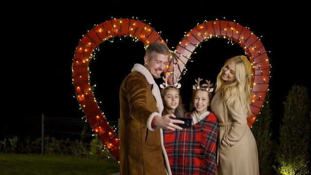 Young family taking a selfie and grimacing, while standing behing heart shaped Christmas decoration. Slow motion. 