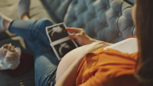 Close-up view of a happy pregnant woman watching her ultrasound report and touching her abdomen while sitting on sofa at home