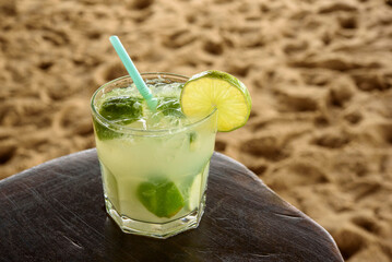 Brazilian caipirinha drink on wooden table with sandy beach background.