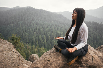 Young woman meditating and breathing on top of a mountain and forest - obrazy, fototapety, plakaty