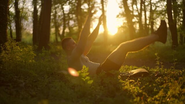 Strong athletic shirtless man doing abdominal exercise on yoga mat in the sunny forest. Healthy lifestyle concept. Slow motion.