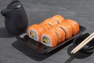 Sushi rolls with salmon, avocado and cucumber on a black rectangular plate on a black stone (slate) background.