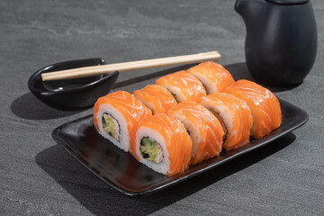 Sushi rolls with salmon, avocado and cucumber on a black rectangular plate on a black stone (slate) background.