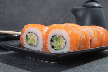 Sushi rolls with salmon, avocado and cucumber on a black rectangular plate on a black stone (slate) background.