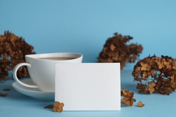 Dried hortensia flowers, sheet of paper and cup with coffee on light blue background. Space for text