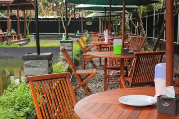 Fototapeta na wymiar Beautiful and simple outdoor seating area beside the fish pond with industrial wooden tables and chairs and big umbrellas above. Tropical plant decoration for outdoor cafe or restaurant