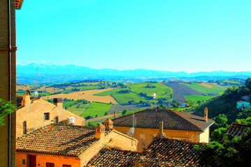 Riveting scenery in Montelupone with different houses and buildings in the foreground, the serene green fields of Marche hilly landscape behind them, and the Sibillini mountains in the background