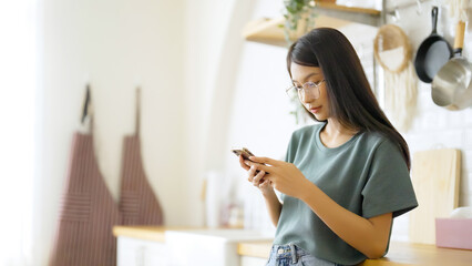 Happy young asian woman wearing glasses and relaxing at home. Asia female standing at counter kitchen and using mobile smartphone
