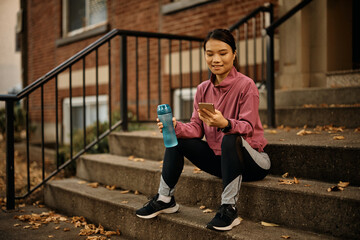 Smiling Asian athletic woman using mobile phone during water break outdoors.