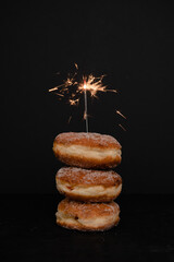 Traditional German or Austrian Neujahrs-Krapfen, doughnut with sparkler fire cracker served for New Year's Eve party celebrations, donut filled with jam and dusted with sugar on black background - 550161006