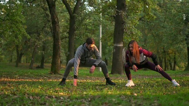 Young beautiful woman and handsome man doing leg stretching after workout in the sunny park.
