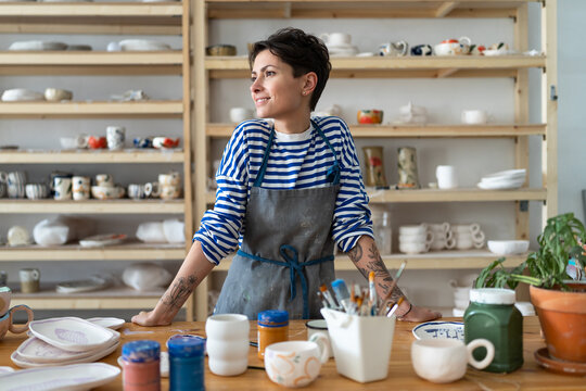 Calm young female owner in pottery workshop looking at window and smiling, plans projects. Small business concept, ceramics studio. Successful italian woman entrepreneur with tattoos at workplace.