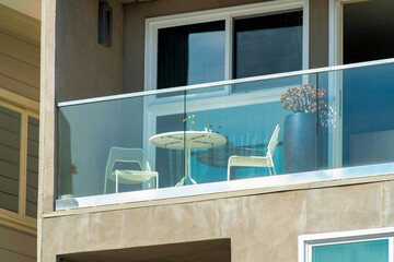 Quaint outdoor balcony with white patio furniture and glass panneling on side of beige stucco building in sun midday