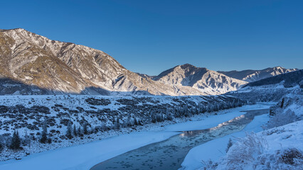 Obraz premium An ice-free river between icy shores. Melted ice and reflection on blue water. Trees and bushes on the snowy shores. The picturesque mountain range is illuminated by the sun against the blue sky.
