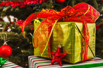 Wrapped Christmas present in gold wrapping paper with a large bow under a decorated Christmas tree
