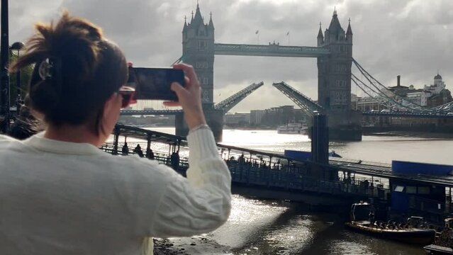 Woman taking a photo with her smartphone of Tower Bridge with open bascules and Thames River in London UK