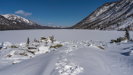 A chain of footprints in snowdrifts goes to the frozen lake. Boulders all around. The surface is...