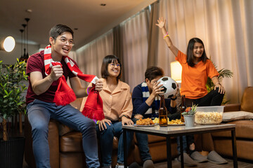 Group of Asian people friends sit on sofa watching and cheering football or soccer games competition on TV together at home.Happy man and woman sport fans celebrating sport team victory sports match
