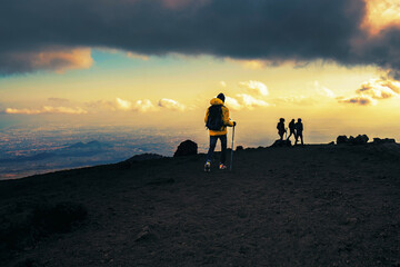 Group of hikers walking through the lava stone of the mount Etna in Sicily, Italy at sunset -...