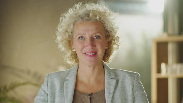 Portrait of a mature blonde haired woman with curly hair sitting at desk, looking at camera and smiling
