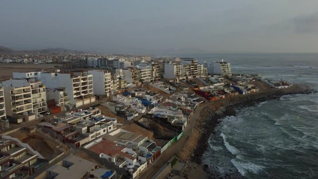 Beach town near the edge of ocean coast. Drone flies back and ascends in altitude revealing buildings into the horizon. Located in Punta Hermosa beach in Lima, Peru.