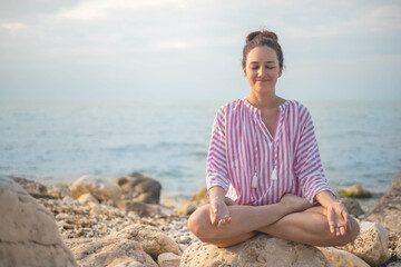 Naklejka na ściany i meble Happy relaxed woman meditation in lotus position practicing yoga sitting on stone beach sea waves