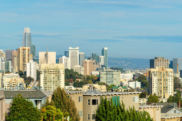 Obraz premium Sprawling city with downtown buildings and structures in evening sun with trees and foliage and foggy blue sky with moutains