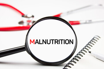 The word malnutrition through a magnifying glass on the background of a stethoscope. Problem of malnutrition concept. - obrazy, fototapety, plakaty