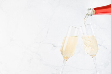 Festive background with flow of pouring fizz champagne from red neck bottle to two glasses on soft light white marble backdrop, copy space, closeup, detail. New Year and celebrate event background.