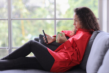 Smart and active latin woman sitting on sofa and using internet on computer with smart mobile phone while drinking coffee in living room. Lifestyle with technology