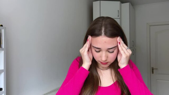 Woman with headache A young girl is holding her head massaging her temples headache closes her eyes and raises her head up holding hands behind pink jacket with long brown hair