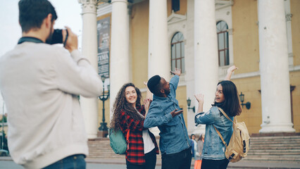 Obraz premium Joyful tourists men and women are posing for camera with beautiful building in background having fun laughing and jumping while bearded guy is holding camera and shooting.