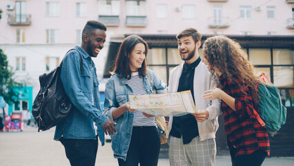 Multiracial group of tourists is looking at paper map standing in the street in modern city talking...