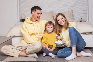 Laughing smiling parents with child son sitting by the bed and eating popcorn