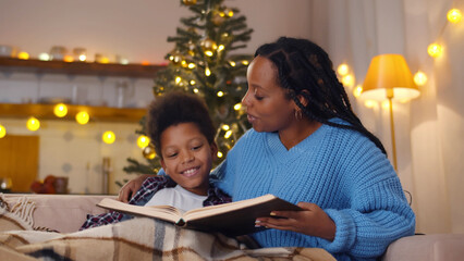 African-American mother and son resting on couch reading book in living room decorated for christmas