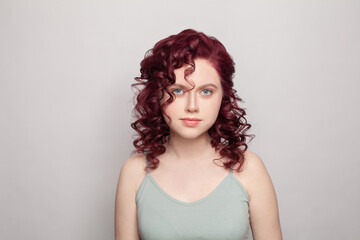 Attractive woman with red curly hair in grey top looking to the camera on white studio wall background