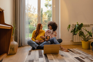 Multiracial couple of two lesbian women sitting on the floor with their baby ultrasound making a video call. Pregnant young redheaded woman
