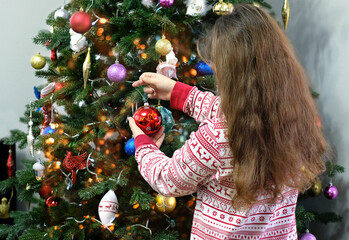 Girl in pajamas holds glass ball next to a decorated Christmas tree. symbol of festive winter...