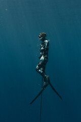 Speardiver swimming to surface with spear in one hand wearing camouflage wetsuit, water is deep blue and clear with sun rays shining through around freediver swimming to surface in Bahamas.
