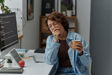 Young tired businesswoman yawning and drinking coffee while sitting at her workplace at office
