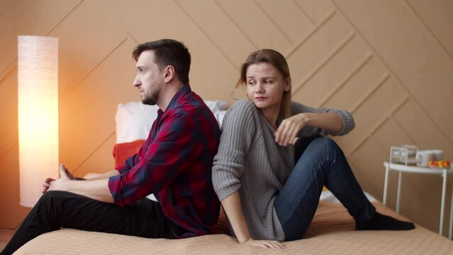Offended man sitting on the bed back to back with the woman, who trying to apologize and putting her hands on man's shoulder, but man does not accept apologies and angrily throwing off her hand