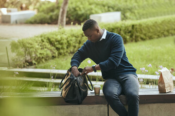 Businessman city park, food and lunch break with coffee or tea drink in paperbag. Young African...