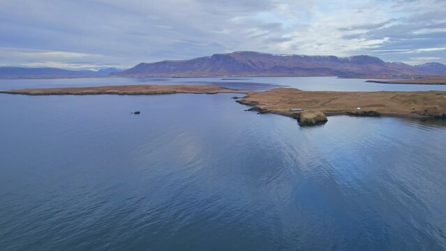 Small Grassy Island Surrounded By Calm Atlantic Ocean Water Surface With Arctic Mountain Landscape In The Background. Aerial Drone Perspective.