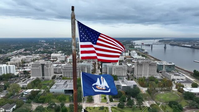 USA and Louisiana state flag wave in breeze. Aerial from atop state capitol building. View of downtown Baton Rouge and Mississippi River.