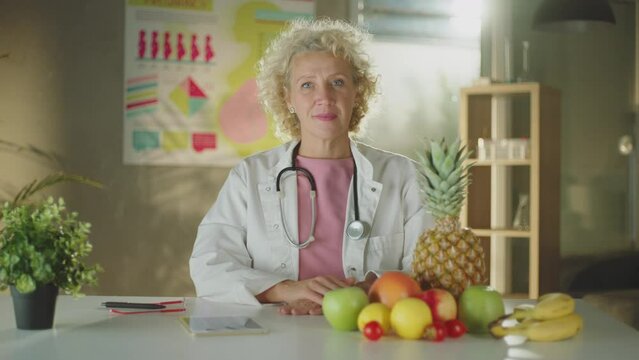 A female doctor sitting with different types of fruits on the desk whiling looking and smiling at camera